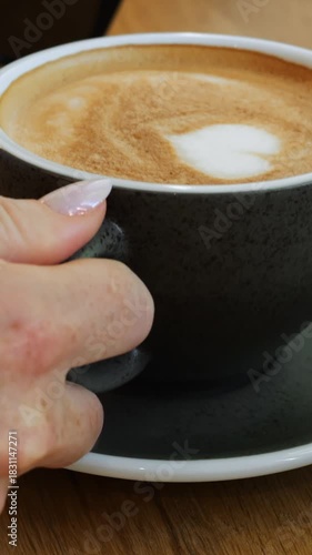 Elegant woman hand takes black ceramic coffee cup from wooden table. Lady guest drinks tasty cappuccino from mug with heart decor in coffee shop vertical video