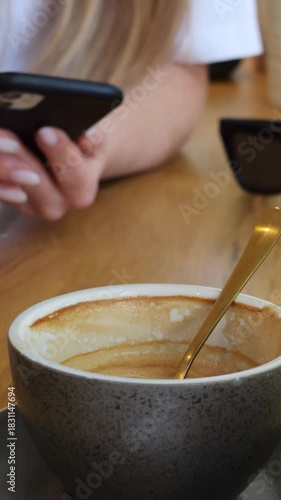 Empty coffee cup on wooden table against woman typing text on smartphone in cafe. Lady chats online via mobile phone during lunch in cozy coffee shop vertical video
