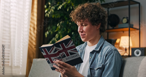 A focused boy studies language at home, holding an English dictionary and reviewing new vocabulary in his thoughts.