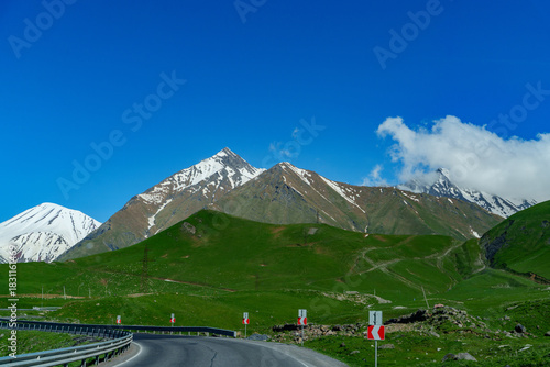 Mountain pass road curving toward bright green slopes and snowy peaks