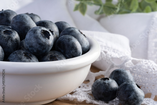 Close-up of fresh blueberries in a white ceramic bowl on a rustic wooden surface with a lace napkin. Bright natural light, minimal food still life with shallow depth of field and copy space