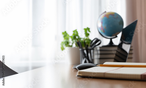 Notebook and pencil on a desk with blurred home office elements in the background — laptop, mouse, books, globe and plant. Cozy remote work, studying or teaching from home concept.