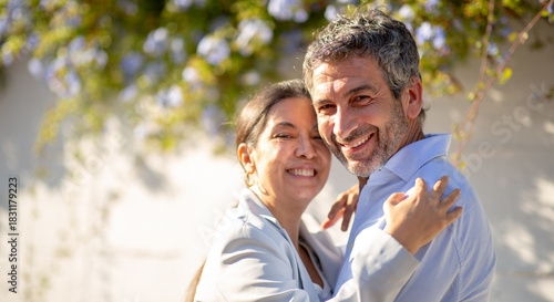 Happy couple embracing and smiling under sunny day in front of a white wall with flowers