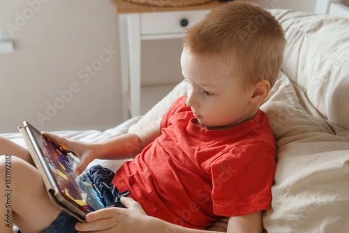Little boy using a digital tablet while relaxing on a bed at home. Kid playing mobile game or watching video. Concept of child screen time.