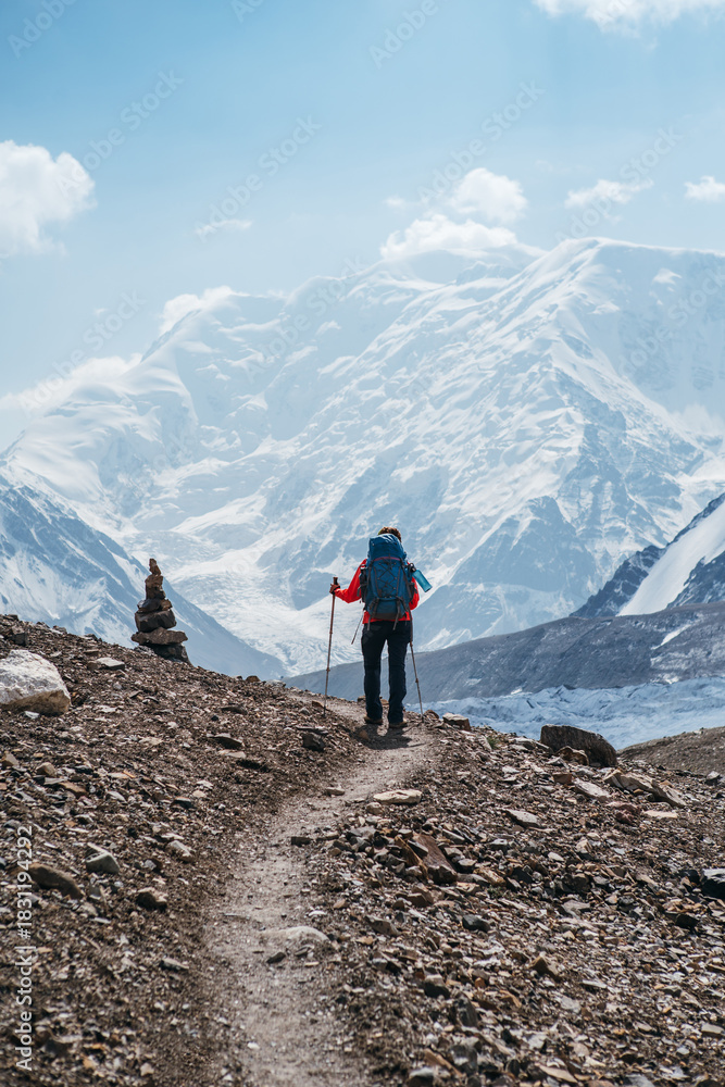 Fototapeta premium Lonely Climber woman with backpack and trekking poles during Lenin peak ascent with mountain peaks and glaciers in background. Extreme active people, high-altitude mountaineering concept image