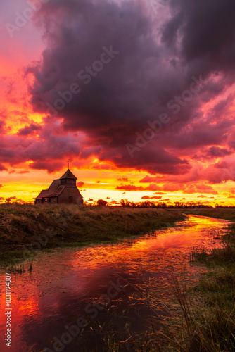 The iconic St Thomas à Becket Church on Fairfield, Kent stands silhouetted against a blazing sunrise, dramatic clouds rolling across the Romney Marsh landscape.
