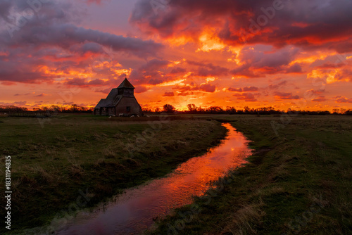 The iconic St Thomas à Becket Church on Fairfield, Kent stands silhouetted against a blazing sunrise, dramatic clouds rolling across the Romney Marsh landscape.
