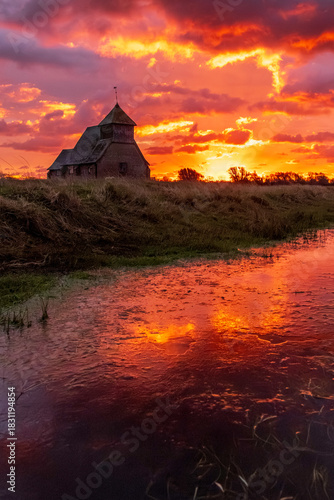 The iconic St Thomas à Becket Church on Fairfield, Kent stands silhouetted against a blazing sunrise, dramatic clouds rolling across the Romney Marsh landscape.