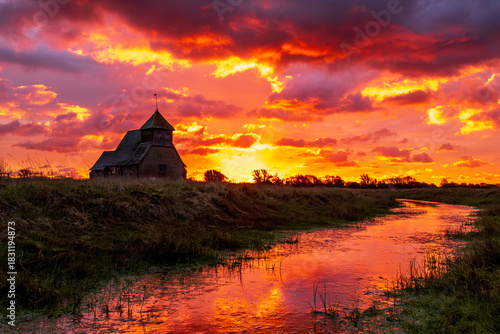 The iconic St Thomas à Becket Church on Fairfield, Kent stands silhouetted against a blazing sunrise, dramatic clouds rolling across the Romney Marsh landscape.