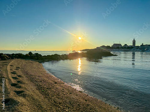 Fototapeta Naklejka Na Ścianę i Meble -  Golden sun rises over the calm Adriatic Sea, casting warm rays on the rocky coastline and illuminating the distant silhouette of Rab town during a peaceful and scenic summer morning view.