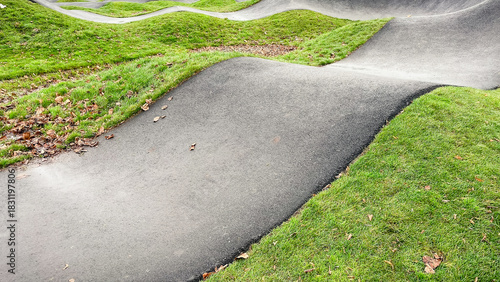 Curved asphalt pump track for cycling and wheeled sports, surrounded by vibrant green grass in a modern park