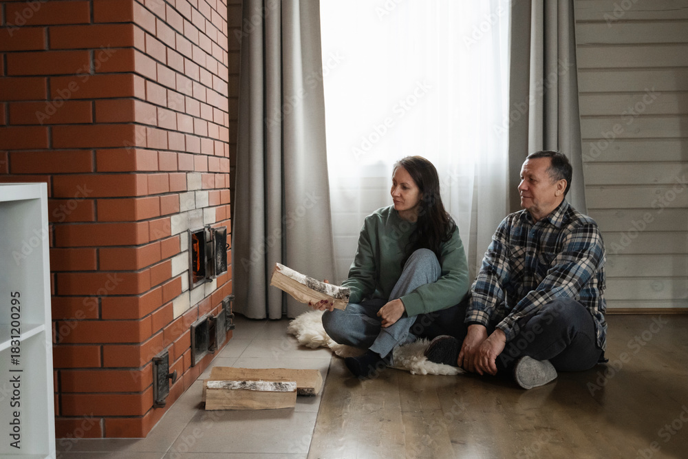 Fototapeta premium Adding firewood to heat stove by caucasian man and woman sitting together on floor in warm indoor rustic room with natural light. Winter Firewood Routine Preparing and Lighting a Wood Stove