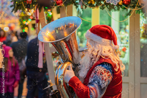 Santa Claus playing tuba at outdoor Christmas market with festive decorations and falling snow. Concept of holiday celebration, music and joyful spirit of Christmas tradition