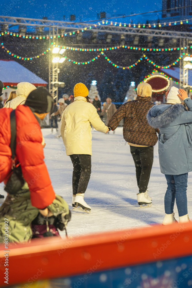 Obraz premium People ice skating on festive illuminated rink under colorful string lights during snowfall. Concept of holiday spirit, cheerful winter entertainment and magical seasonal atmosphere