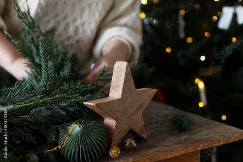 Making Christmas wreath. Hands arranging wreath with fir and cedar branches, and scissors, rustic star on wooden table on background of stylish xmas tree. Winter holidays preparation and decor