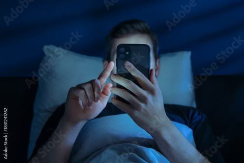 Obraz na plátně Close up of young man, alone in the dark bedroom, holds his smartphone close to face while scrolling social media or texting late at night