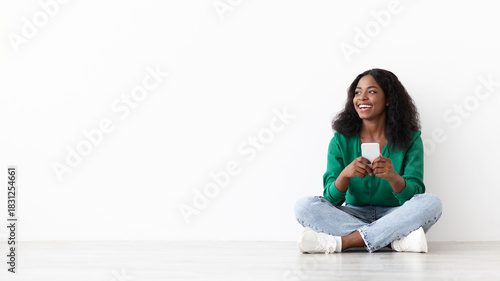 A young woman sits on the floor with her legs crossed and holds a smartphone, smiling with joy in a well-lit environment. Her casual outfit includes a green top and jeans.