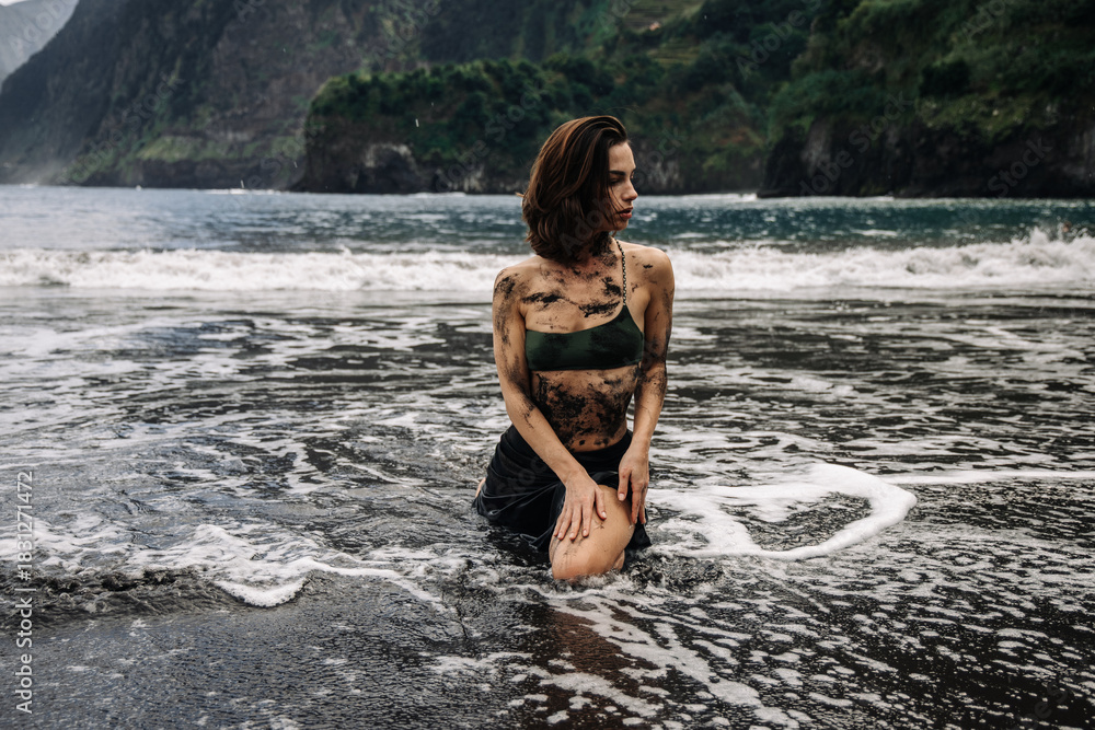 Fototapeta premium Young woman with wet hair kneeling in ocean waves on a rocky beach, wearing a green bikini, surrounded by lush cliffs and dramatic coastal scenery, embodying freedom and nature
