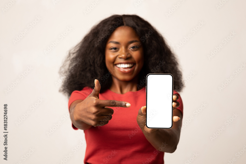 Fototapeta premium A cheerful Black woman shows excitement while pointing at her smartphone with a blank white screen. She stands against a light background, perfect for showcasing app ads.