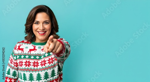 Happy young adult woman in her 30s wearing a festive Christmas sweater with reindeer and trees, smiling and pointing directly at the camera on a vibrant blue studio background