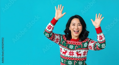 Happy young Asian woman in a festive red and green Christmas sweater with reindeer and snowflakes, smiling joyfully with arms raised on a vibrant blue background, celebrating the holiday.