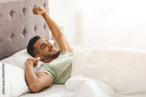 Obraz na plátně A young man sits up in bed, stretching his arms and smiling in a bright, sunny bedroom