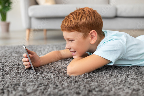 Cuadro en lienzo A smiling redheaded preteen boy is watching a video on his mobile phone while lying on the carpet