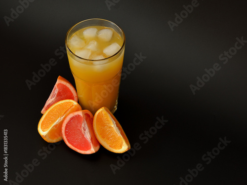 Fruit juice with ice on a black background in a tall glass next to slices of ripe orange and grapefruit.