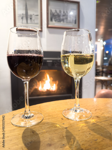 Two glasses of wine one white one red on a table in front of a warm cosy winter fireplace with orange flames