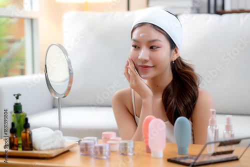 Beauty young asian woman applying cream on facial for wrinkles while looking mirror in living room at home, happy beautiful woman applying lotion moisturizer on cheek for hygiene, skincare and beauty.