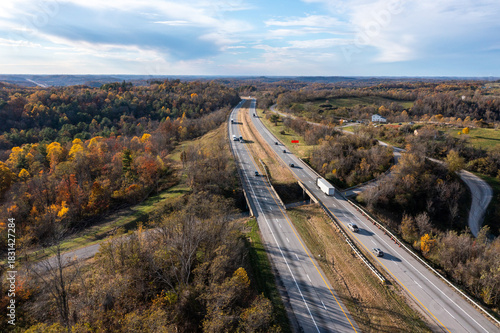 
aerial view of a highway cutting through colorful autumn hills, showing moving cars, soft light, and wide open landscape. peaceful fall scenery captured from above.
