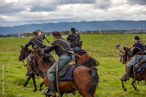 Civil war reenactment scene with union cavalry riders charging across an open field. horses in motion, historical uniforms, and dramatic cloudy mountains in the background create an action-filled mome