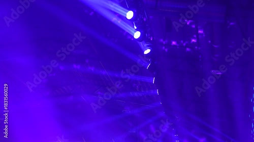 Concert lighting rig generating dramatic beams and atmospheric color patterns as moving head fixtures cast vivid blue and purple illumination across the venue during a professional stage show