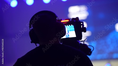 Event videographer filming stage performance with professional camera rig, wearing headphones and monitoring live feed in blue illuminated venue environment during production