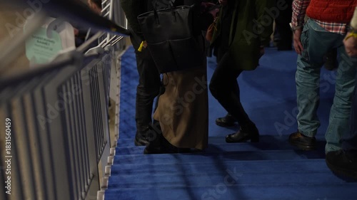 People walking down a blue carpeted staircase inside an event venue during crowd movement, showing legs, shoes and clothing as attendees exit the area in an indoor environment