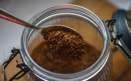 A spoon dipped into a glass jar filled with instant coffee granules on a wooden table, captured in warm natural light.