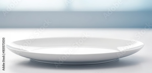 A minimalist empty white ceramic plate positioned on a light reflective surface with a soft blue background