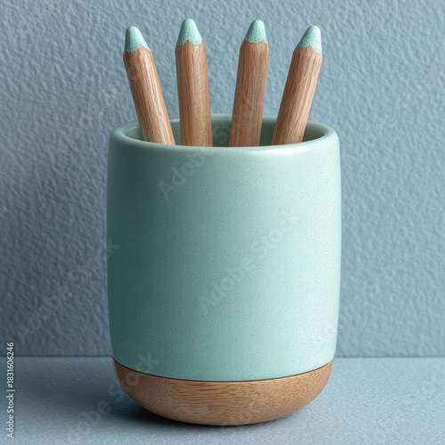 A close up studio shot of four wooden pencils with mint green tips contained within a speckled mint green ceramic holder with a wooden base, resting on a textured gray surface.