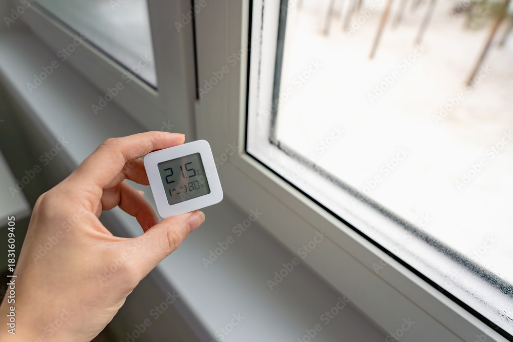 Fototapeta premium High indoor humidity indicated by a digital hygrometer showing 80 percent, next to a wet window with visible condensation in winter, indoor climate control concept.