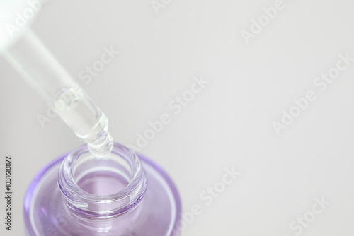 Close-up of a dropper releasing serum into a purple glass bottle on a light background with copy space. Ideal for skincare, cosmetics, beauty products, wellness, and self-care themes.