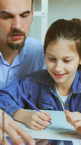 Father points at tablet while daughter writes in notebook, both focused during learning activity at desk in bright indoor space