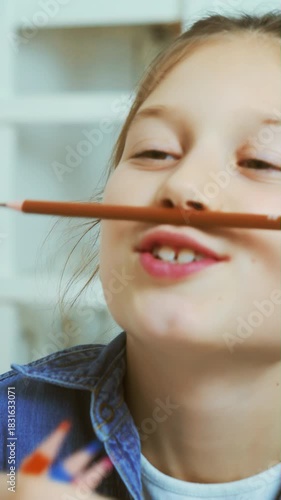 Smiling girl in denim shirt playfully holds pencil under nose while puffing cheeks and making silly face during lighthearted moment indoors