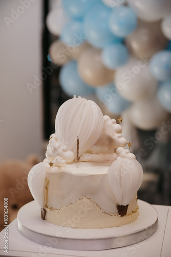 Cake with a candle on top, standing on a stand in the middle of a restaurant against a backdrop of a screen of inflatable colorful balloons