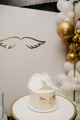 A cake stands on a white table in a restaurant against a white screen with an image of angel wings and inflated balloons