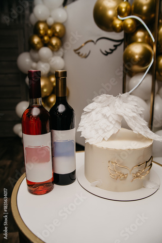 A cake stands next to two bottles of wine on a white table in a restaurant against a white screen.