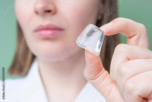 Young woman holding ice cube near her face on green background.