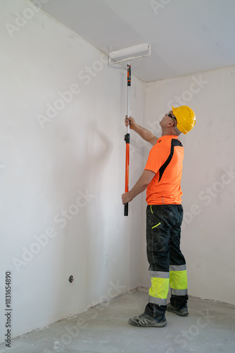 A construction worker paints a ceiling with a wide, specialized paint roller. Apartment renovation.