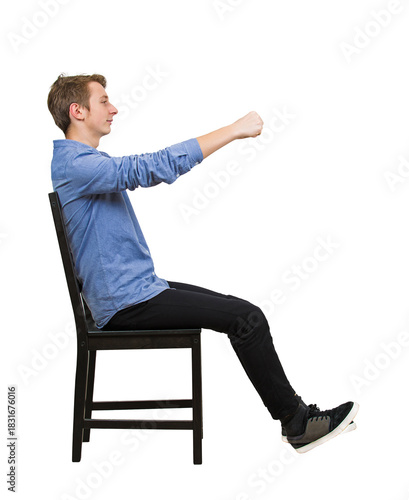 Side view full length portrait of confident teenage guy driver, pretending to drive an imaginary transportation, while sitting on a chair with arms stretched as holding the steering wheel