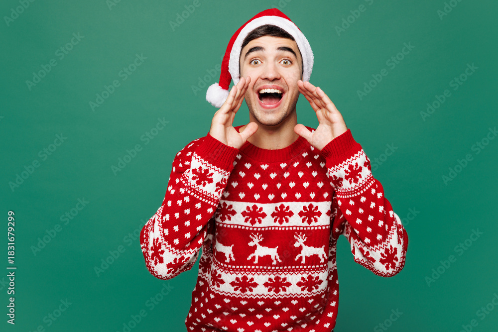 Fototapeta premium Merry young man wear red Christmas sweater Santa Claus hat posing scream sharing hot news about sales discount with hands near mouth isolated on plain green background. Happy New Year holiday concept.