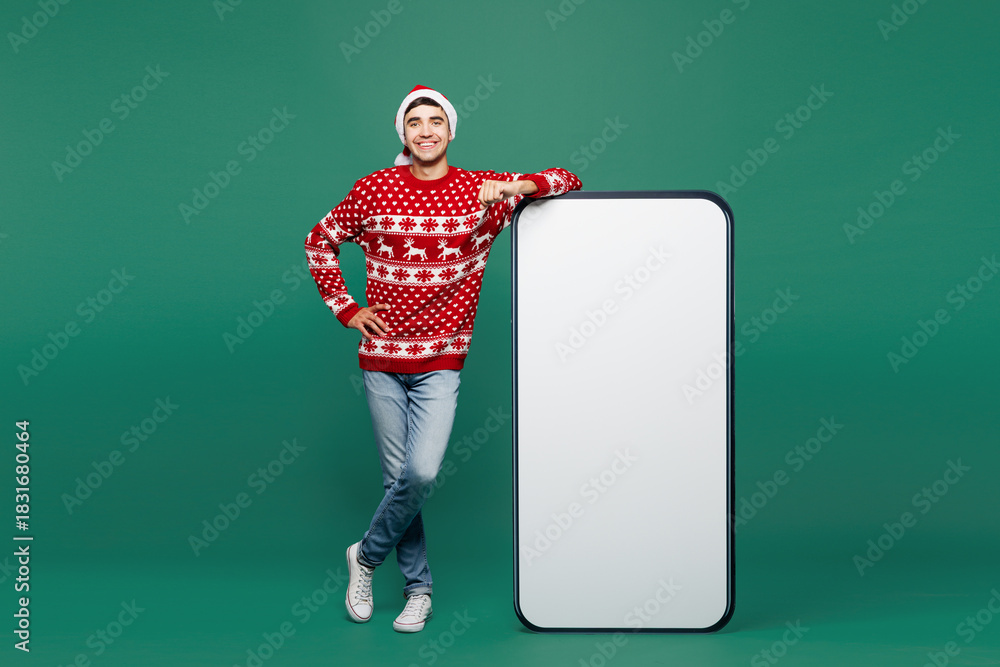 Fototapeta premium Full body merry young man wear red Christmas sweater Santa Claus hat posing next to big huge blank screen mobile cell phone area smartphone isolated on plain green background. Happy New Year concept.